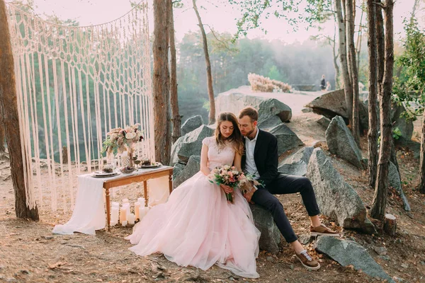 Atractivos pareja de recién nacidos, felices y alegres momentos. Hombre y mujer vestida de fiesta se sientan en las piedras cerca de la decoración de la boda en estilo boho. Ceremonia. — Foto de Stock