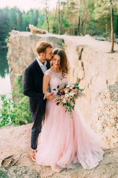 Groom está beijando sua noiva. Felizes recém-casados abraçando. Homem em tuxedo e mulher em um vestido de noiva rosa está posando sobre a natureza. Cerimônia ao ar livre. Close-up — Fotografia de Stock