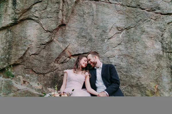 Casal atraente recém-casados noiva e noivo ri e sorri para o outro, feliz e alegre momento. Homem e mulher em roupas de casamento sentar-se no fundo da rocha. — Fotografia de Stock