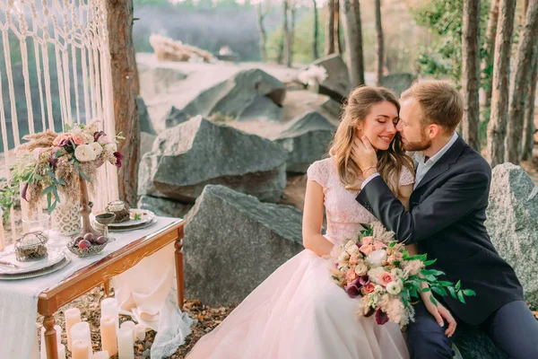 Attractive couple newlyweds, tender moment before a kiss. Man and woman in festive clothes sit on the stones near the wedding decoration in boho style. Stock Photo