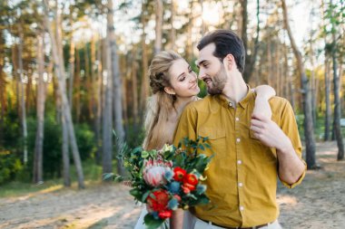 Güzel gelin onun damat tarafından omuz kucaklar. Bir ormandaki düğün yürüyüş. Yeni evli birbirine benziyor. Kız beyaz elbise ve bir zeytin tişörtlü adam. Sanat.
