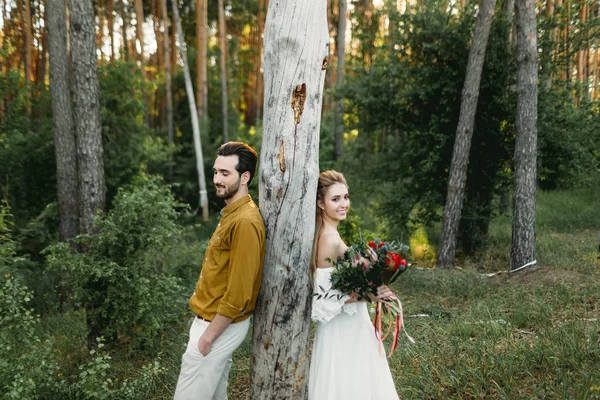 Braut und Bräutigam lehnen sich von verschiedenen Seiten an den Baum. Frischvermählte gehen im Wald spazieren. Kunstwerk — Stockfoto