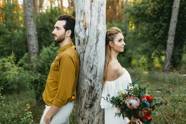 Bride and groom lean on the tree from different sides. Newlyweds are walking in the forest. Artwork — Stock Photo, Image