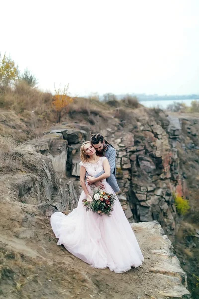 Şefkatle kucaklayan arkasında tarafından ona güzel gelin damat. Sonbahar düğün töreni rustik tarzı açık havada. Yeni evliler Dağları arka plan üzerinde poz olan — Stok fotoğraf