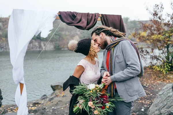 Atractiva pareja recién casados ríen y sonríen momento feliz y alegre. Ceremonia de boda de otoño al aire libre. Novia y novio con estilo con rastas se miran el uno al otro de pie ante un lago — Foto de Stock