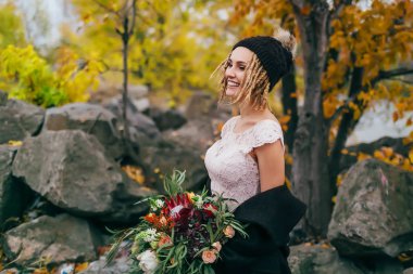 Rustik bir buket ile şık gelin bir göl kenarında tepeden önce poz. Genç sarışın ponpon ve dreadlocks ile örme şapka içinde. Sonbahar düğün töreni açık havada.