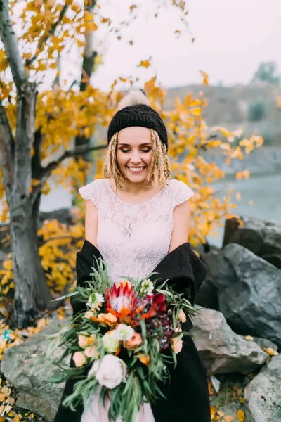 Novia con estilo con un ramo rústico está posando ante un lago en la colina. Joven rubia en un sombrero de punto con pompón y con rastas. Ceremonia de boda de otoño al aire libre . — Foto de Stock