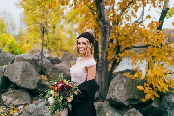 Стильная невеста с деревенским букетом позирует перед озером на холме. Молодая блондинка в вязаной шляпе с помпоном и дредами. Осенняя свадебная церемония на открытом воздухе . — стоковое фото