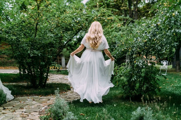 Underbara blonda bruden i vit klänning promenader i den grönskande trädgården. Bakifrån. Konstverk. — Stockfoto
