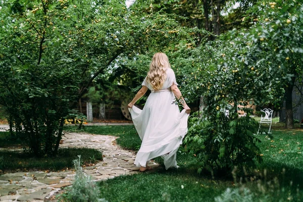 Hermosa novia rubia en vestido blanco está caminando en el jardín verde. Vista trasera. Obra de arte . — Foto de Stock