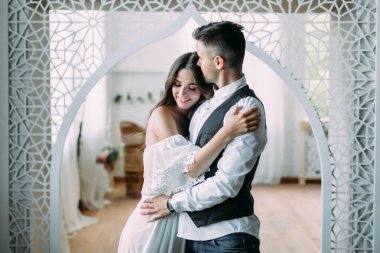 Gülümseyen ve onu kafasından öper ve beline kucaklayan damat sarılma neşeli genç gelin. Vintage odada poz güzel yeni evliler