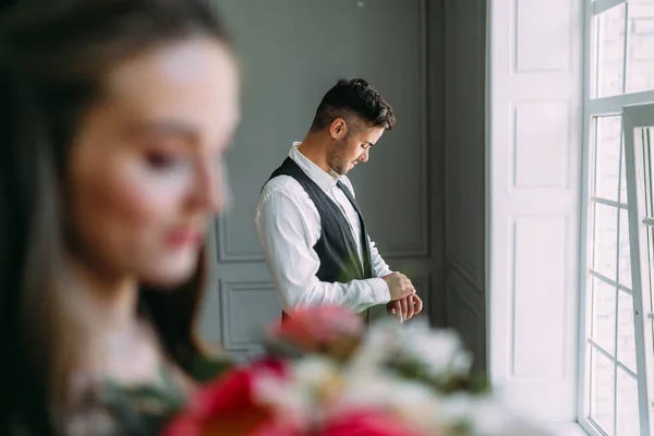 Красивий наречений готується до весільного дня на тлі розмитої нареченої з букетом. Чоловік у святковому одязі біля панорамного вікна. Художня робота — стокове фото