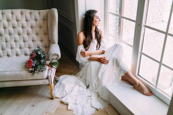 Una mujer junto a la ventana. Novia mirando por la ventana, espera al novio. Un ramo de novia se encuentra en el sofá . — Foto de Stock