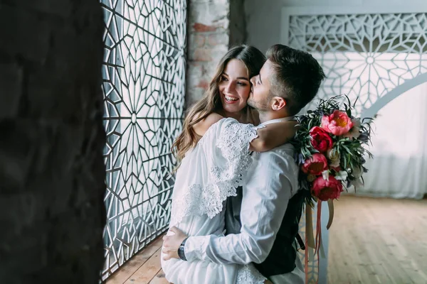 Joven novia alegre abrazando novio mientras él la besa en la mejilla y abraza su cintura en el alféizar de la ventana — Foto de Stock