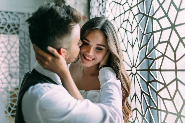 Весела молода наречена обіймає голову наречених, поки він цілує її в щоці на старовинному фоні вікна — стокове фото