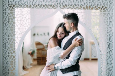 Gülümseyen ve onu kafasından öper ve beline kucaklayan damat sarılma neşeli genç gelin. Vintage odada poz güzel yeni evliler