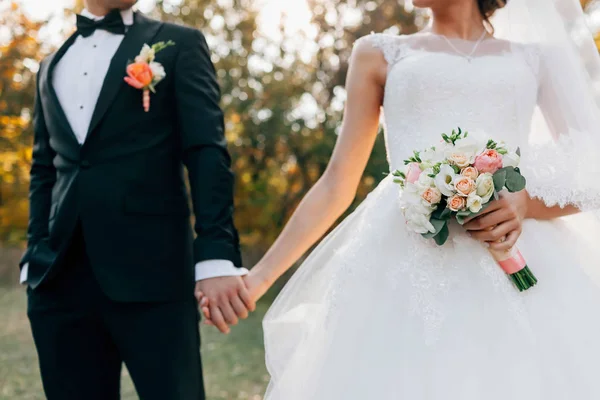 Bouquet de mariage. Mariée floue avec une robe blanche et marié en smoking se tiennent la main. Concentration douce sur les fleurs. À l'extérieur — Photo