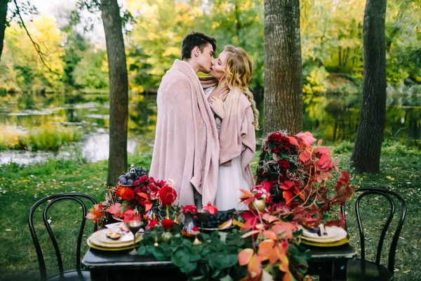 Newlyweds kissing under the plaid next to the festive table. Bride and groom in the park. Autumn wedding. Artwork — Stock Photo, Image