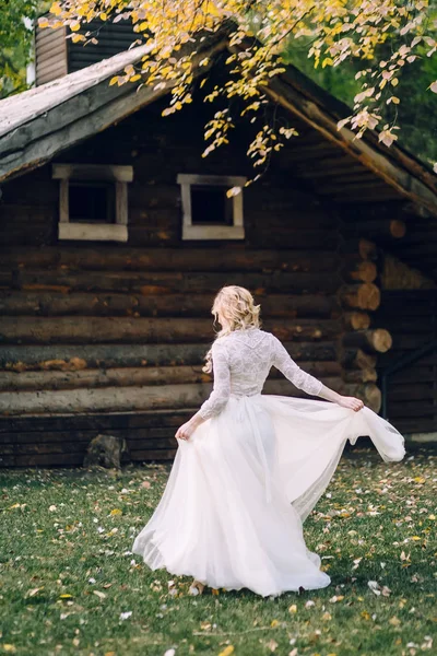 La hermosa novia está girando alrededor de sí misma en vestido revoloteando sobre el fondo de la casa de madera. Vista trasera. Obra de arte — Foto de Stock
