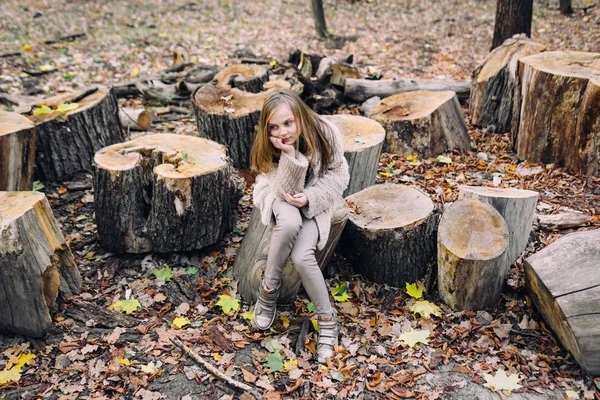 Little girl is sitting on wooden stump in the forest at autumn day. — Stock Photo, Image