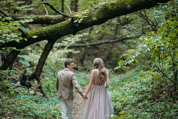 A happy couple is walking on a trail in an autumn forest. Bride and groom are looking at each other on nature. — Stock Photo, Image