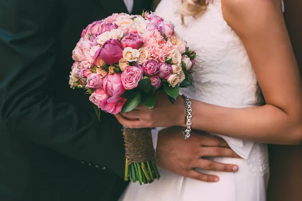 A noiva bonita mantém um buquê de casamento com rosas rosa e peônias. Groom abraçar mulher pela cintura . — Fotografia de Stock