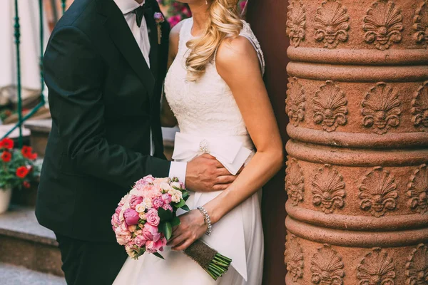 Bella sposa abbronzata in un abito bianco e lo sposo in smoking sono in piedi accanto al muro e in possesso di un bouquet da sposa. Sposo abbracciare la donna per la vita. All'aperto — Foto Stock