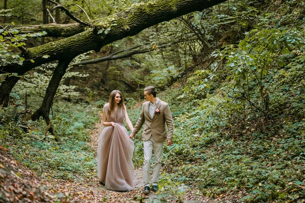 A happy couple is walking on a trail in an autumn forest. Bride and groom are looking at each other on nature. — Stock Photo, Image