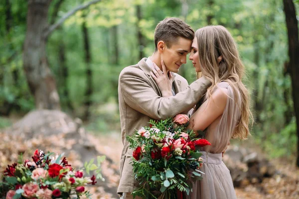 Стильная пара обнимает и трогает лбы, стоя в лесу. Осенняя свадебная церемония на открытом воздухе . — стоковое фото