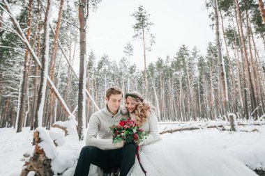 Gelin ve damat bir buket ile oturum açma arka plan karlı ormanın oturuyor. Düğün kış. Sanat