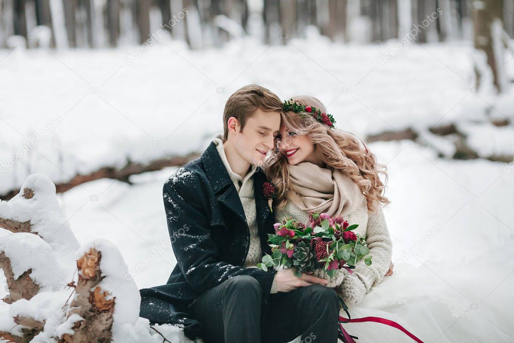 Cute couple in love with a bouquet are sitting on the log on background of the winter forest. Artwork