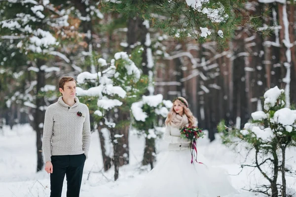 英俊的新郎在米色套衫的背景下, 模糊的新娘在白雪皑皑的森林。冬季婚礼。艺术品. — 图库照片