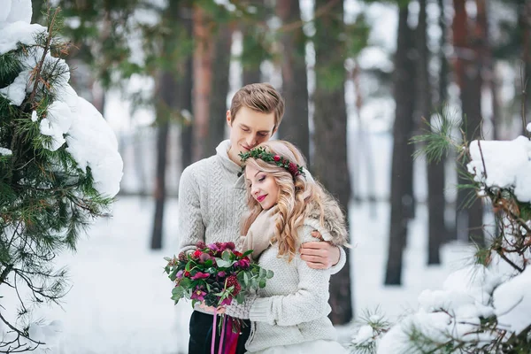 Bride with wreath and bouquet, groom in beige knitted pullover are walking in snowy forest. Winter wedding. Artwork. — Stock Photo, Image