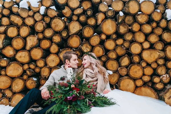 Noiva e noivo olham um para o outro. Os recém-casados elegantes sentam-se na neve no fundo de madeira. Casamento de inverno — Fotografia de Stock