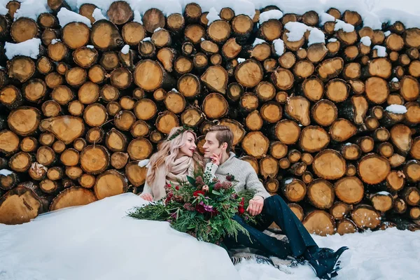 Braut und Bräutigam schauen einander zärtlich an. Stylisches Brautpaar sitzt auf Schnee auf dem hölzernen Hintergrund. Winterhochzeit — Stockfoto