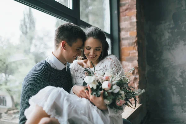 Glada nygifta bredvid fönstret. Brudgummen håller sin fru på händerna hemma. Konstverk — Stockfoto