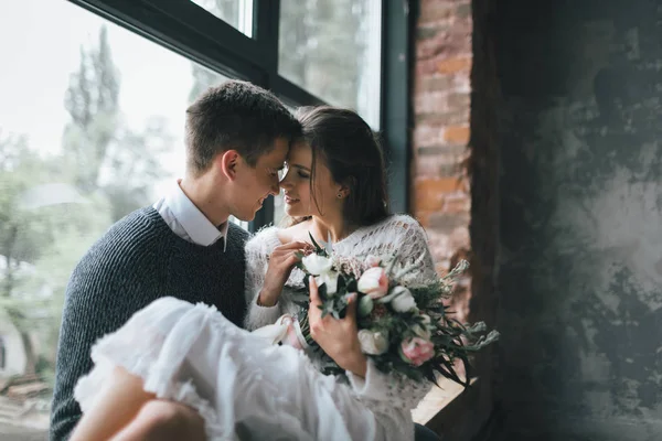 Glada nygifta bredvid fönstret. Brudgummen håller sin fru på händerna hemma. Konstverk — Stockfoto