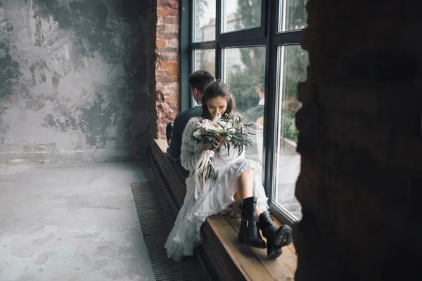 La elegante novia con bouquet se sienta en la ventana junto a la novia. Obras de arte Imagen de stock