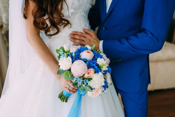 Mariée dans une robe blanche et marié dans un smoking bleu sont debout à côté de la fenêtre et tenant un bouquet de mariage . — Photo