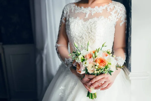 Una hermosa novia está de pie cerca de la ventana y la celebración de un ramo de bodas con rosas blancas y peonías de melocotón. Primer plano —  Fotos de Stock
