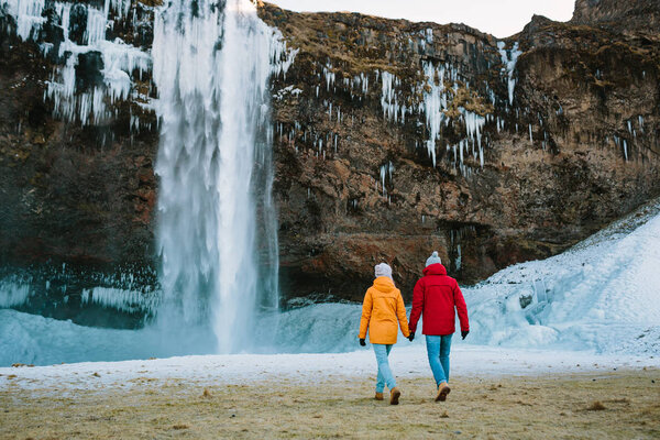 Man in red jacket and woman in yellow jacket walking together next to waterfall. Iceland travelers