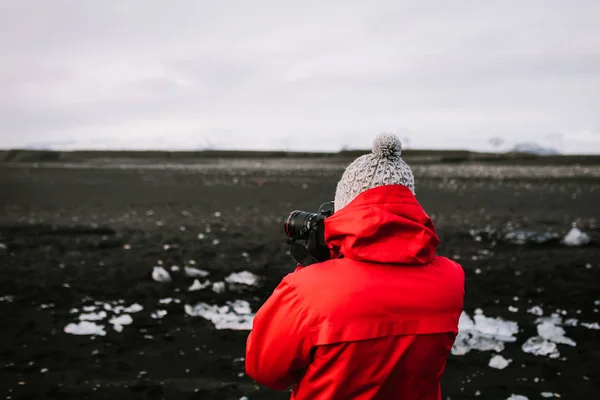 Fotógrafo en chaqueta roja tomando una foto de la playa negra Islandia naturaleza. Vista trasera —  Fotos de Stock