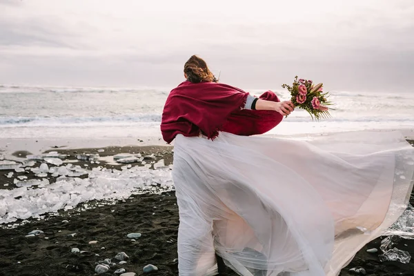 Невеста в белом льстивом платье с букетом прогулок в ветреный день на Черном песке Бич, Исландия. Произведения искусства. Ее волосы трясутся от ветра — стоковое фото