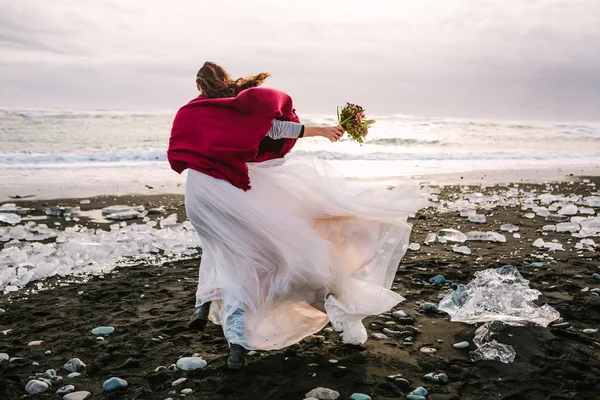 Невеста в белом льстивом платье с клеткой бургундской прогулки в ветреный день на Черном песке Бич, Исландия. Произведения искусства. Ее волосы трясутся от ветра — стоковое фото