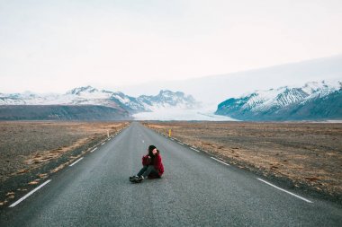 Kırsal güzel dağları arka plan üzerinde de yol oturan genç hipster kadın. Sıcak giysiler içinde bir kız. Rüzgarlı hava.