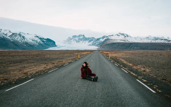 Happy young girl sit on the middle of road on background of beautiful mountains. She is in warm clothes. Windy weather. — Stock Photo, Image