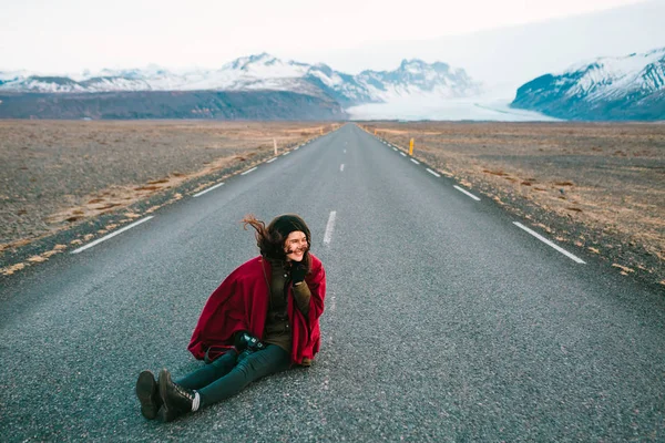 Young cheerful woman sitting on the road with photocamera on background of beautiful mountains. Iceland tourist. Windy weather. — Stock Photo, Image