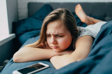 Üzgün genç kadın yatakta yalnız başına uzanmış, üzgün beyaz kadın kendini yalnız hissediyor çünkü mesajcıdaki arkadaşları tarafından cevaplanmamış yataktaki sorunu düşünüyor.