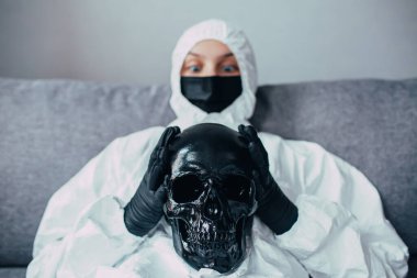 Siyah bir maske takmış, viral koruyucu giysi ve siyah tıbbi eldiven giymiş, elinde siyah bir kafatası tutan, ona bakan, Coronavirus yüzünden sonuçlardan ve ölümden korkan paniğe kapılmış bir hemşire.
