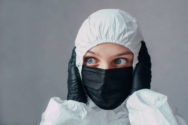 Viral koruyucu giysili, tıbbi maskeli ve siyah eldivenli bir doktor elleri başının üzerinde pencereye korku ile bakıyor. Virolog, koronavirüs salgınının sonuçlarından korkuyor.